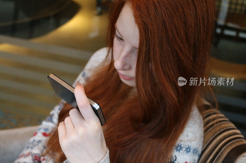 一个14 / 15岁的红发少女，皮肤苍白，脸上有雀斑，坐在室内专注地盯着手机屏幕